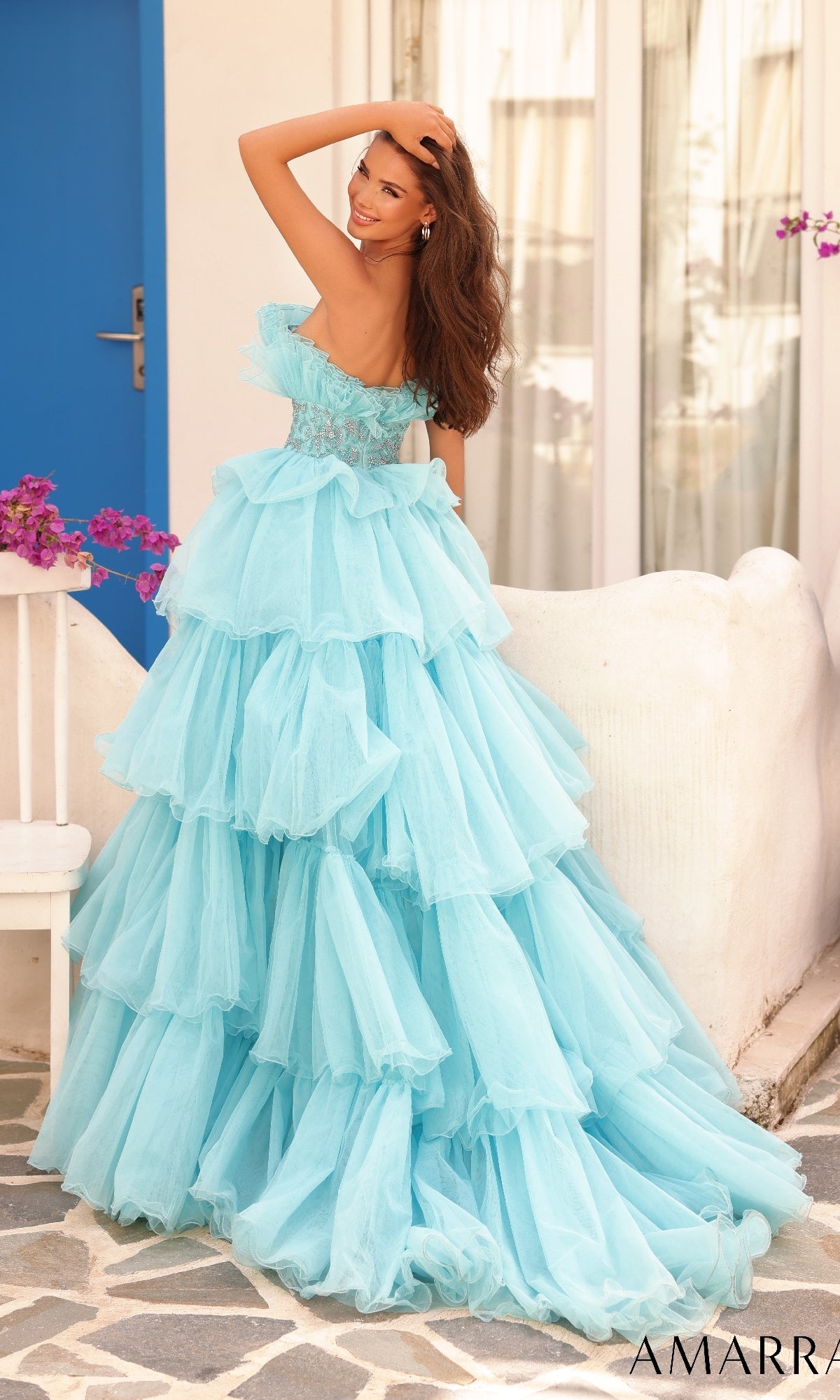 A woman poses outdoors on a stone pathway near white walls and a blue door, wearing the Long Formal Dress 88083 by Amarra?a strapless gown with a beaded waist?her hand gently touching her hair.